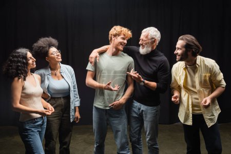 happy producer talking to redhead man near smiling interracial actors on theater stage