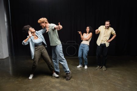Foto de Cheerful interracial students pointing at redhead man and african american woman rehearsing on theater stage - Imagen libre de derechos