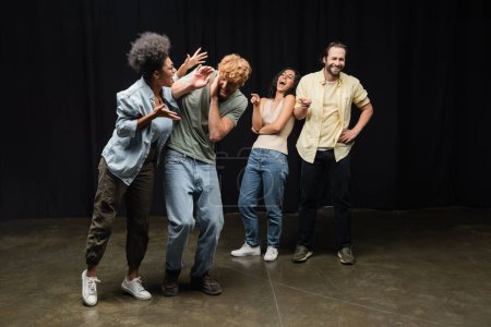 cheerful interracial actors pointing at african american woman rehearsing with redhead man on stage in theater