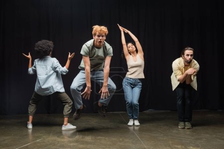 excited redhead man jumping near multiethnic actors posing during rehearsal in theater