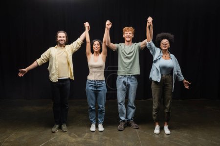 full length of young and joyful interracial actors holding raised hands and smiling at camera in theater