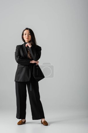full length of brunette and asian businesswoman in black suit standing on grey 