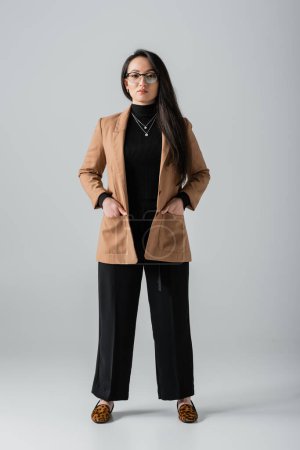 full length of young asian businesswoman in beige blazer and black pants standing with hands in pockets on grey 