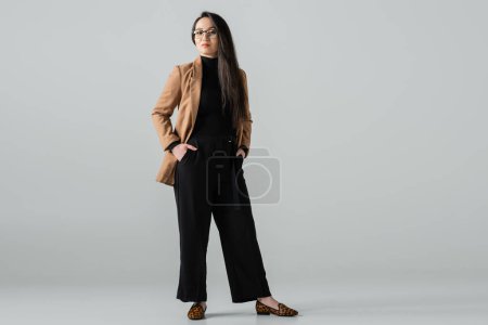 full length of confident asian woman in beige blazer and black pants standing with hands in pockets on grey background 