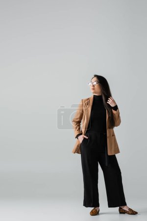 full length of confident asian businesswoman in beige blazer and black pants standing with hand in pocket on grey 
