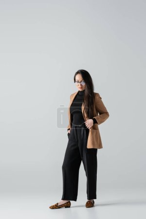 Photo for Full length of asian businesswoman in beige blazer and black pants standing with hand in pocket on grey - Royalty Free Image