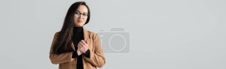 Photo for Young asian businesswoman in glasses and beige blazer standing with clenched hands isolated on grey, banner - Royalty Free Image