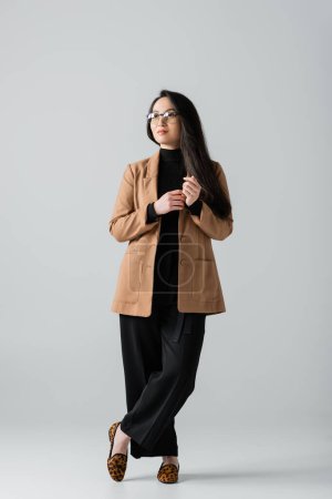Photo for Full length of young asian businesswoman in beige blazer and black pants standing with crossed legs on grey - Royalty Free Image
