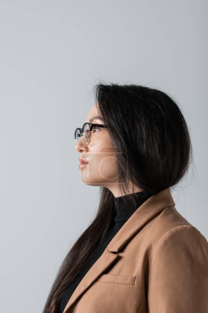 Foto de Side view of young asian businesswoman in glasses and beige blazer looking away isolated on grey - Imagen libre de derechos