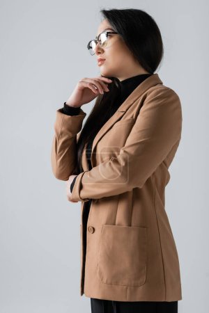 young asian woman in glasses and blazer looking away isolated on grey 