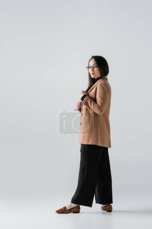 full length of young asian woman in beige blazer and black pants posing on grey 