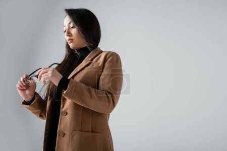 low angle view of asian businesswoman in beige blazer and black turtleneck looking at glasses isolated on grey 