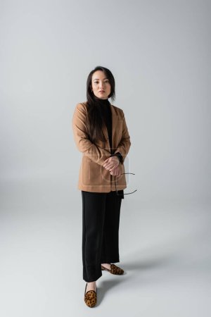 full length of young asian woman in beige blazer and black pants holding glasses on grey 