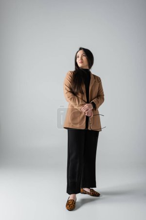 full length of brunette asian woman in beige blazer and black pants holding glasses on grey 