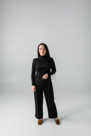 full length of young asian woman in black pants and turtleneck standing on grey 