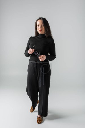 full length of asian woman in black outfit and loafers with animal print holding glasses on grey 