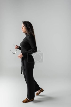 full length of brunette asian woman in black outfit and loafers with animal print holding glasses on grey 