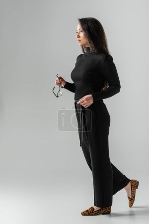 full length of brunette asian woman in stylish black outfit and loafers with animal print holding glasses on grey 