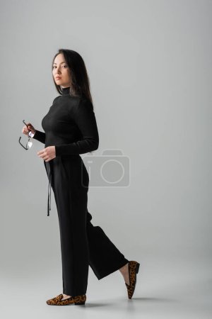 full length of brunette asian woman in stylish black outfit and loafers standing with glasses on grey 