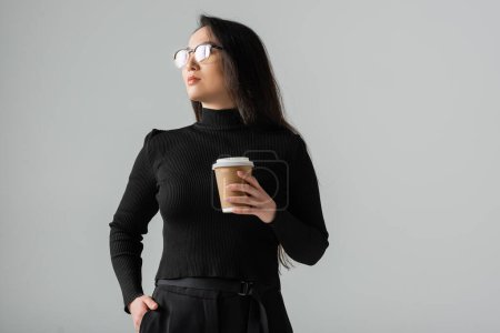 brunette asian woman in stylish black turtleneck holding paper cup while posing with hand in pocket isolated on grey 