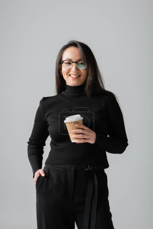 happy asian woman in stylish black turtleneck holding paper cup while posing with hand in pocket isolated on grey 