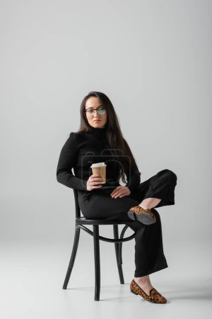 full length of asian woman in black outfit and glasses holding paper cup while sitting on chair on grey 