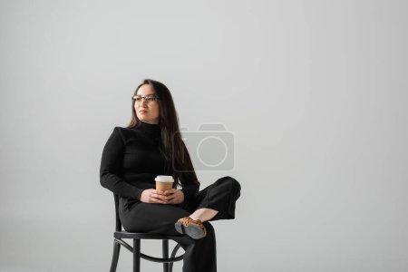 brunette asian woman in black outfit and glasses holding paper cup while sitting on chair isolated on grey 