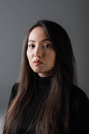 portrait of pretty and asian woman in black turtleneck looking at camera isolated on dark grey 