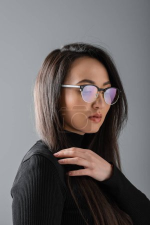 portrait of brunette and asian woman in black turtleneck and glasses isolated on dark grey 