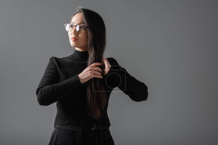portrait of brunette and asian woman in black turtleneck and glasses looking away isolated on dark grey 