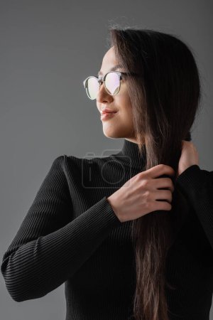 smiling asian woman in black turtleneck and glasses looking away while touching hair isolated on dark grey 