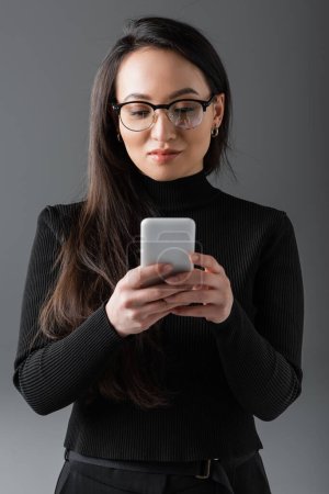 pretty asian woman in black turtleneck and glasses texting on smartphone isolated on dark grey 