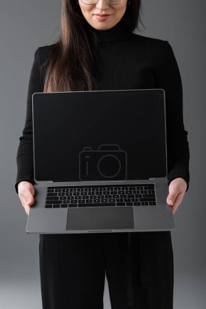 cropped view of smiling woman in black turtleneck showing laptop with blank screen isolated on dark grey 