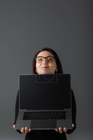 pleased asian woman in black turtleneck and glasses holding laptop with blank screen isolated on dark grey 
