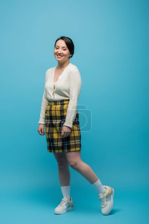 full length of cheerful asian woman in checkered skirt and sneakers standing on blue 