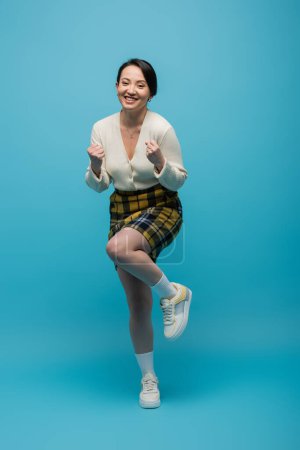 full length of positive asian woman in checkered skirt and sneakers standing on blue 