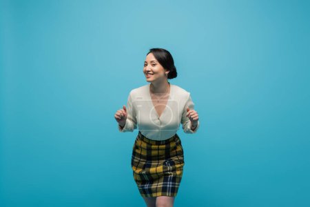 cheerful asian woman in plaid skirt smiling while posing isolated on blue 