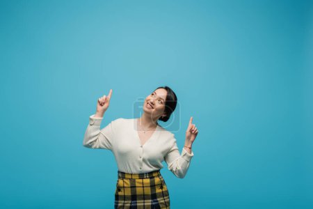cheerful asian woman in plaid skirt smiling while pointing up with fingers isolated on blue 
