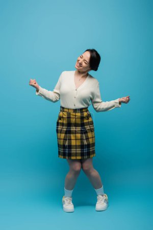 full length of joyful asian woman in checkered skirt and white sneakers standing on blue 