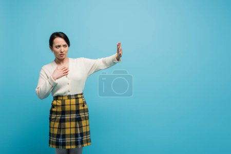 young asian woman in plaid skirt standing with outstretched hand while showing stop isolated on blue 