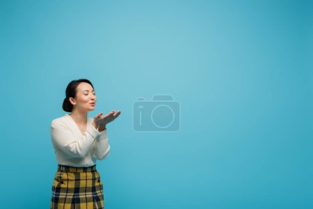 young asian woman with closed eyes sending air kiss isolated on blue 
