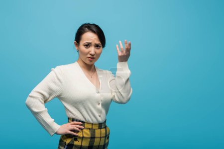 Photo for Confused asian woman in cardigan looking at camera isolated on blue - Royalty Free Image