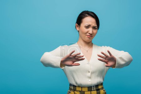 Confused asian woman showing stop gesture isolated on blue 