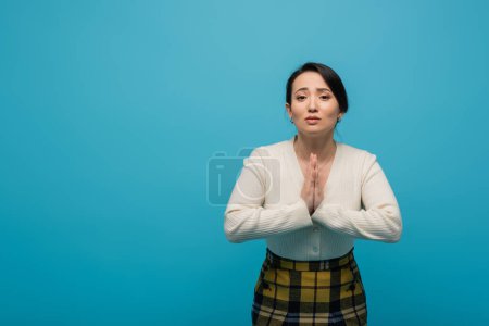 Foto de Asian woman in cardigan showing praying hands gesture isolated on blue - Imagen libre de derechos