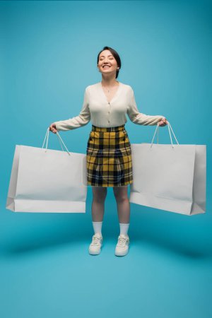 Positive asian woman holding shopping bags while standing on blue background