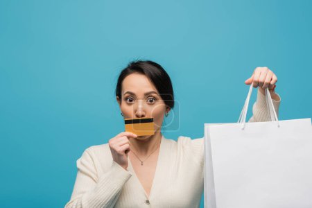 Young asian woman holding credit card near face and shopping bags isolated on blue 