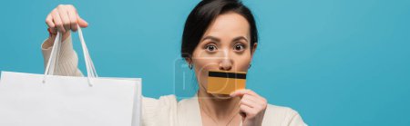 Brunette asian woman holding shopping bags and credit card near face isolated on blue, banner 
