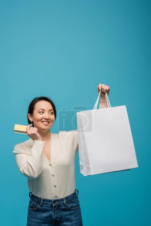Pleased asian woman holding credit card and looking at shopping bags isolated on blue 