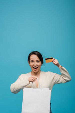 Excited asian woman in cardigan holding shopping bags and credit card isolated on blue 