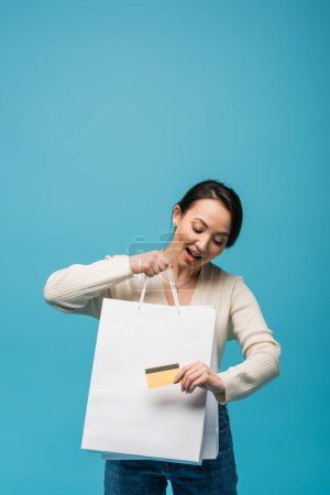 Astonished asian model holding credit card near shopping bags isolated on blue 